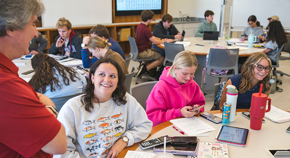 A teacher talk to a table of students in a packed classroom.