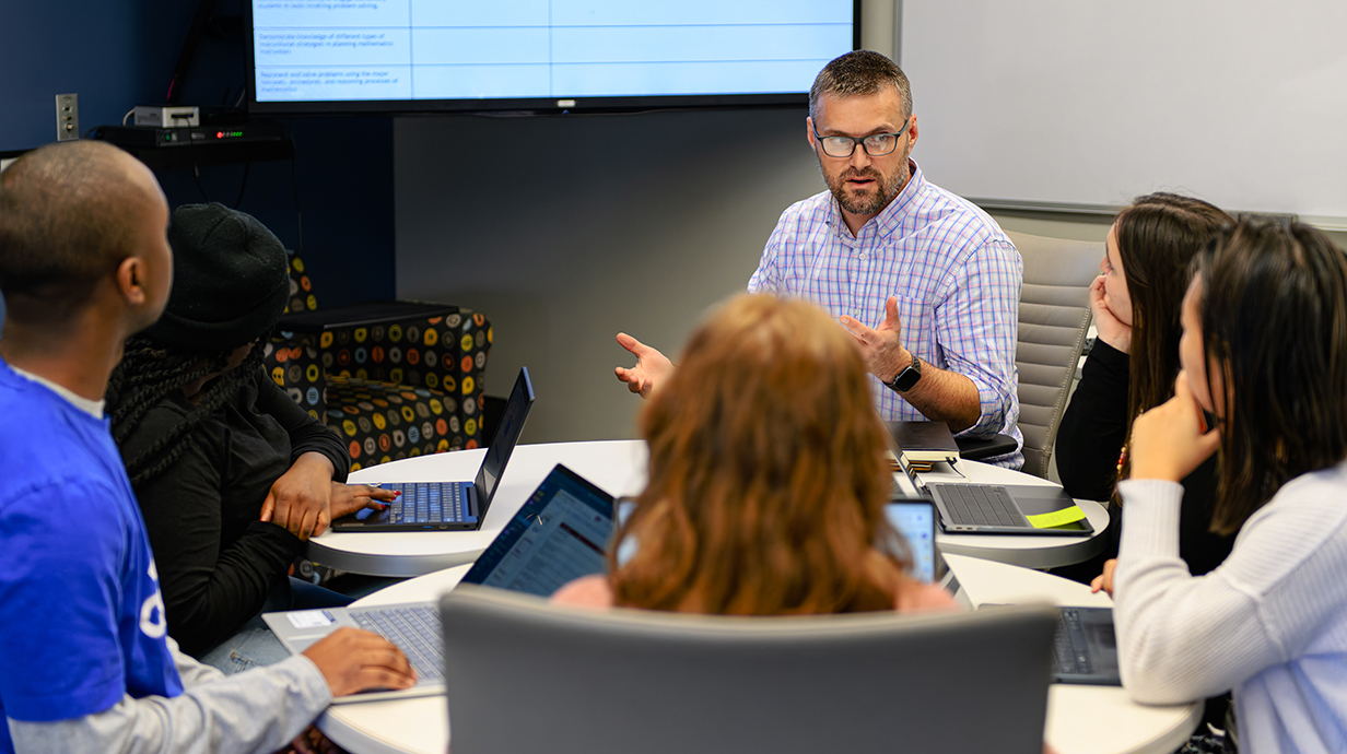 A man talks to several young people seated around a round table in a classroom.